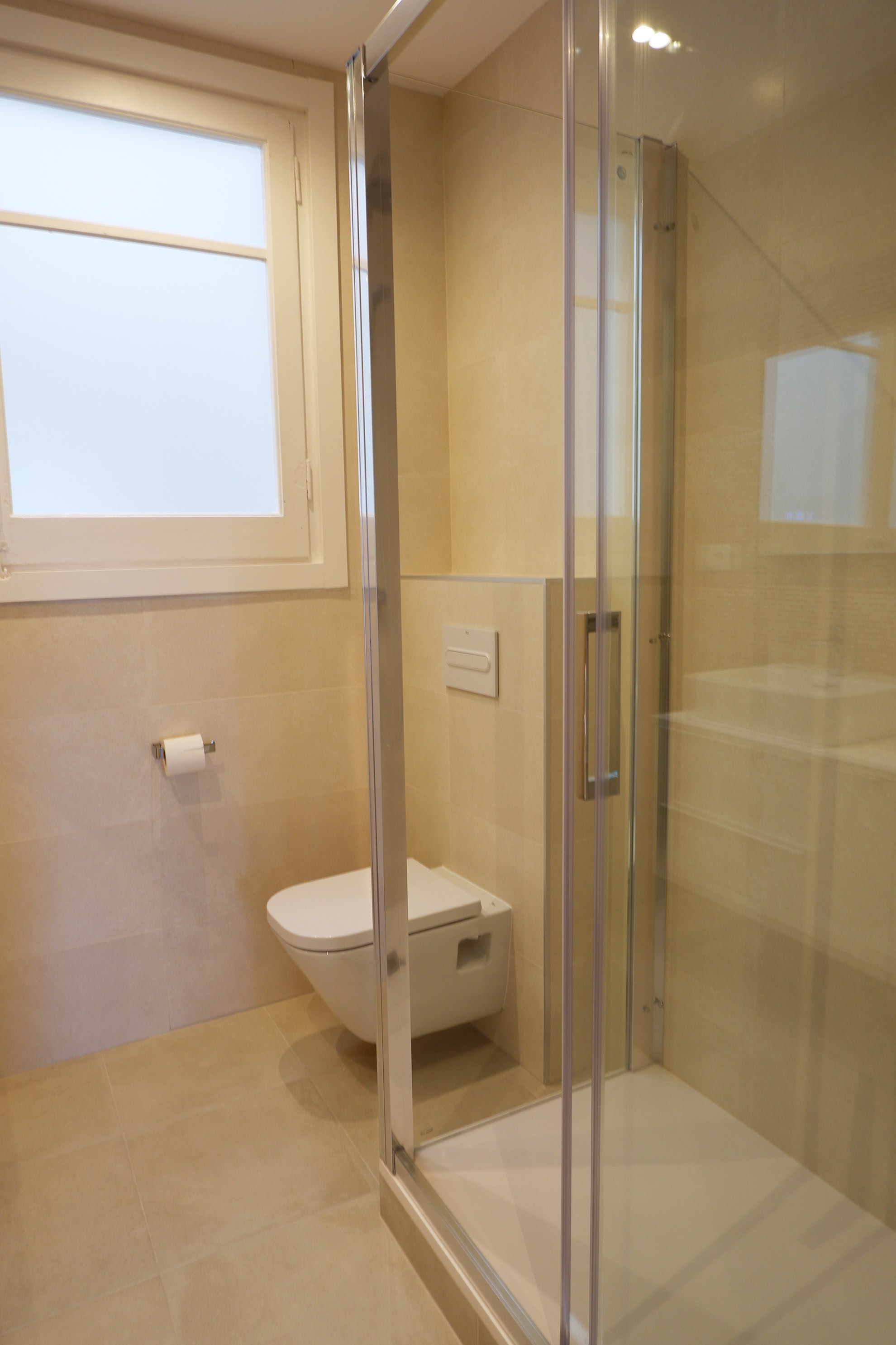Reforma de baño principal en l'Eixample con baldosa beige clara de gran formato, inodoro Roca suspendido con cisterna empotrada y diseño moderno en Barcelona.